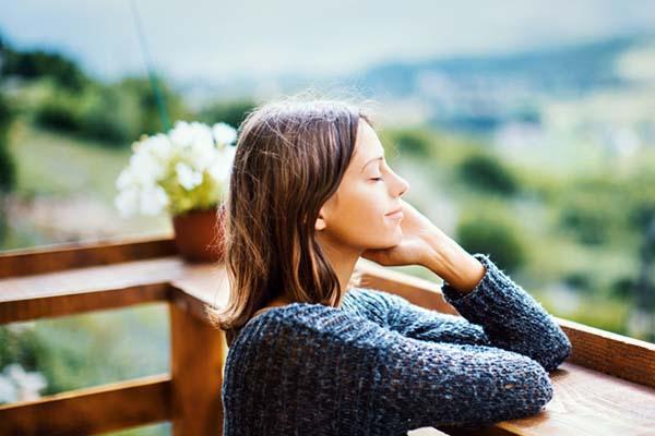 A young woman stands on a deck outdoors with her eyes closed, appreciating the natural surroundings