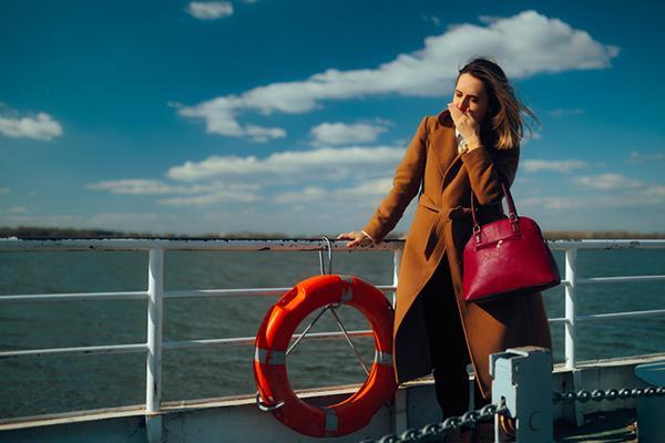 A woman standing on the deck of a ship experiencing motion sickness: she is covering her mouth with one hand and gripping the rail with the other.