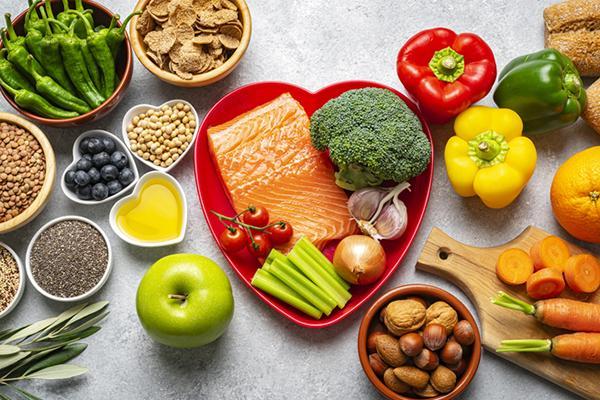 Food assortment viewed from above, centered on a red heart-shaped plate holding salmon and broccoli, surrounded by multicolored peppers, whole-grain breads, dishes of nuts and seeds, berries, oil, and an apple.