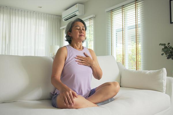 A woman sits cross-legged on a couch with a hand on her chest, engaged in a breathing exercise.