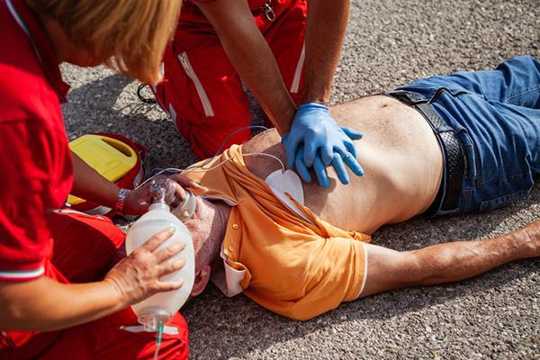 Emergency responders perform CPR on an unconscious man lying on asphalt. A paramedic provides chest compressions while another administers oxygen, with defibrillator pads attached to the man's chest.