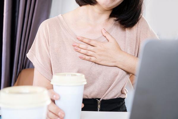 Cropped image shows the torso of a woman holding a coffee cup in one hand, with the other on her chest as she experiences a rapid heartbeat.