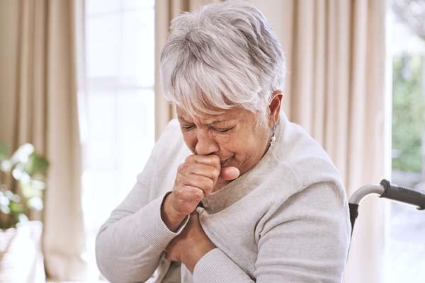 A senior woman sitting in a wheelchair coughs, holding a closed hand in front of her mouth.