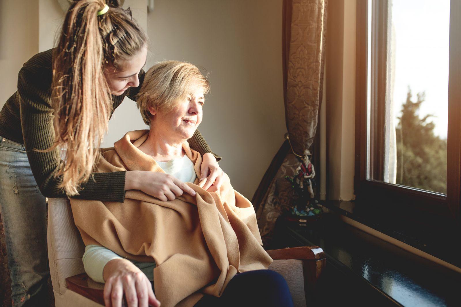 Older woman sitting in a chair and staring out the window with a younger woman leaning over her shoulders