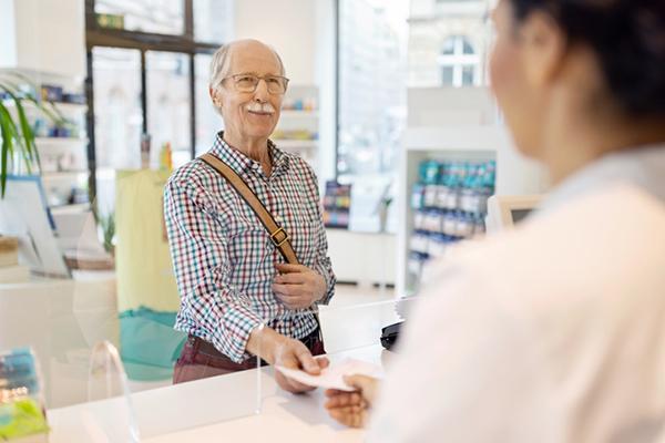 A senior man smiles as he hands a written prescription to a pharmacist; the view is from behind the pharmacist, over the shoulder.