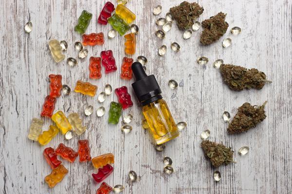 Overhead view of a glass bottle containing CBD oil lying on a wooden table surrounded by dried marijuana buds, CBD extract capsules, and CBD gummies in assorted colors.