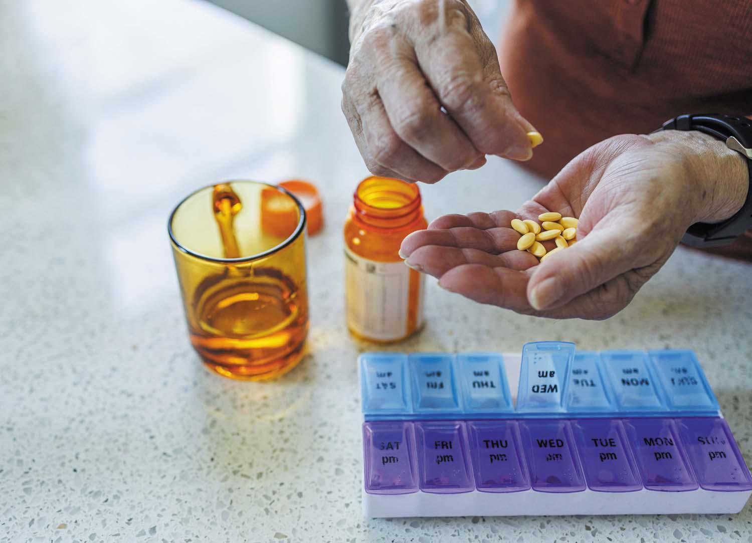 cropped photo showing the hands of a person filling a weekly prescription pill box with yellow pills held in their palm; an orange medication bottle is open in front of them next to a glass of water