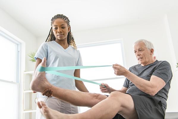 A physical therapist watches as a senior man performs leg exercises using a resistance band.