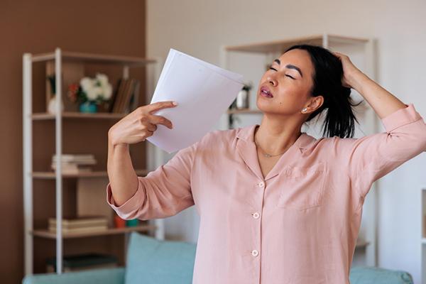 A mature woman in menopause experiencing a hot flash, fanning herself with paper in one hand while holding back her hair with the other.