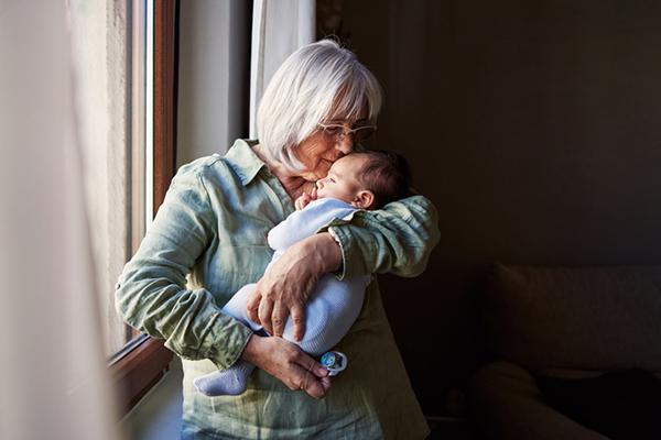 A senior woman looks happy as she holds her grandson while standing next to a window.