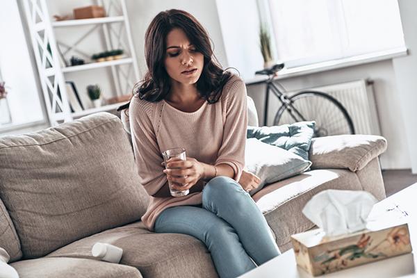 A woman experiencing stomach discomfort sits on a sofa with a glass of water in one hand and the other holding her stomach.
