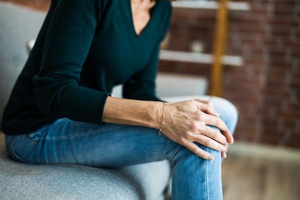 Cropped image of the torso and legs of a woman holding both hands on her knee, experiencing pain.