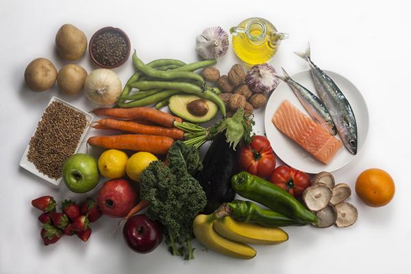 An assortment of healthy foods including fish, olive oil, green beans, carrots, broccoli, garlic, nuts and seeds, apples, strawberries, eggplant, and more.