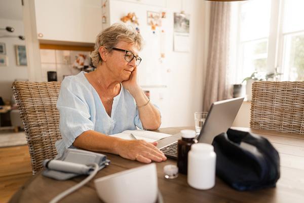 A senior woman sits at her kitchen table, using a laptop to engage in a telemedicine visit.