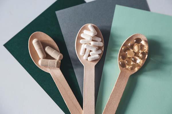Overhead view of three wood spoons holding three different varieties of vitamin capsules; the spoons are positioned on colored paper in dark green, gray, and light green.