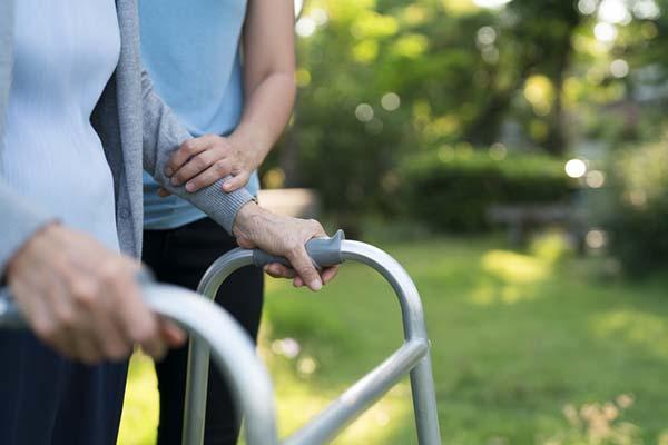 Cropped image of the midsections of two women, one is being helped to walk using a walker by a caregiver, outdoors with trees in the background, out of focus.