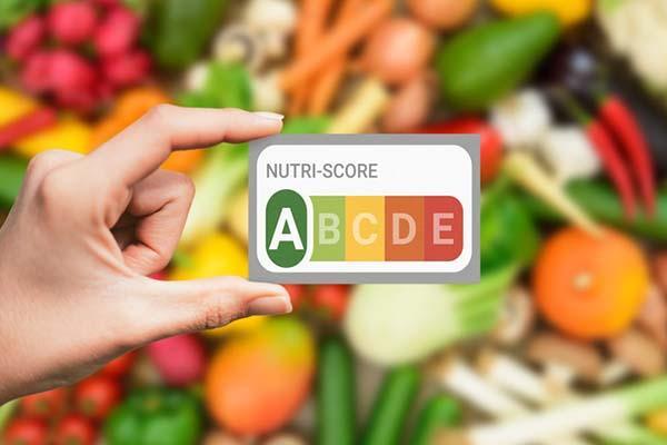 A hand holding a Nutri-Score card indicating nutritional value from a to E, with a colorful assortment of healthy, colorful vegetables in the background, out of focus.
