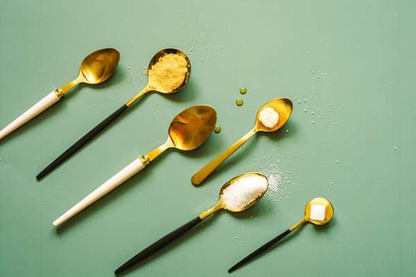 Assorted types of sugar on gold-colored spoons with either wihte or black handles, arranged in parallel on a green surface.