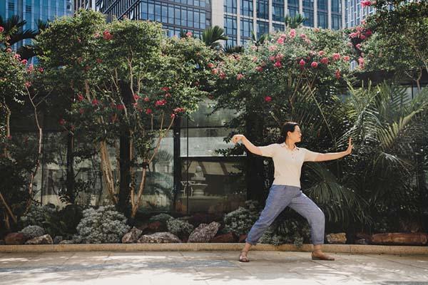 A woman holds a tai chi pose in an ubran setting; immediately behind her are flowering trees with buildings visible behind them.