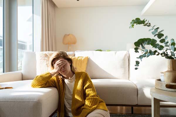 A woman sits on the floor in front of her couch, holding a hand to her forehead and looking distressed.