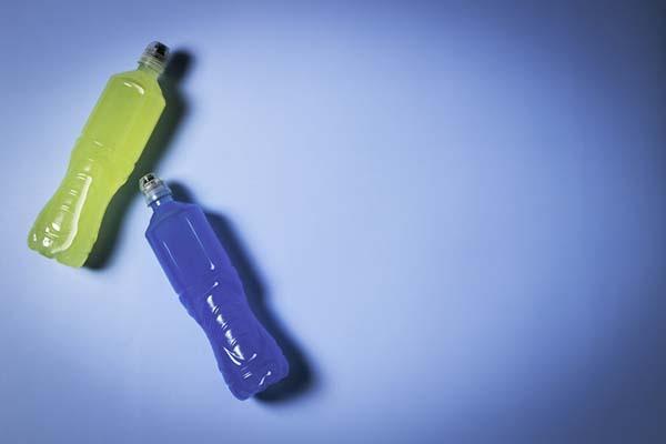 Two plastic bottles filled with energy drinks, one yellow-green and one blue, lying on their sides on a lighter blue surface.