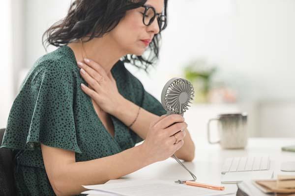 A woman sitting at a desk experiencing a hot flash, holding a small powered fan in one hand.