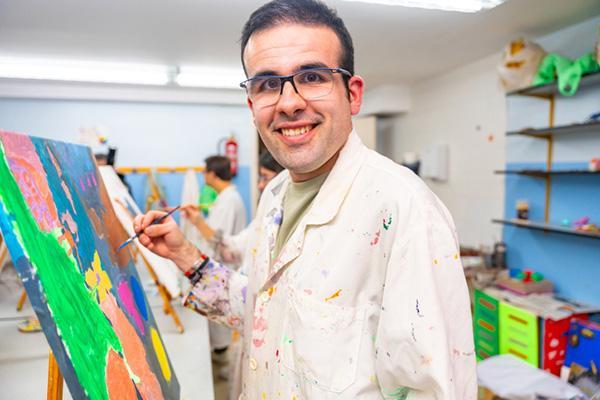 A man wearing glasses smiling and looking at the camera, wearing a smock and standing next to an easel with a painting he is working on.