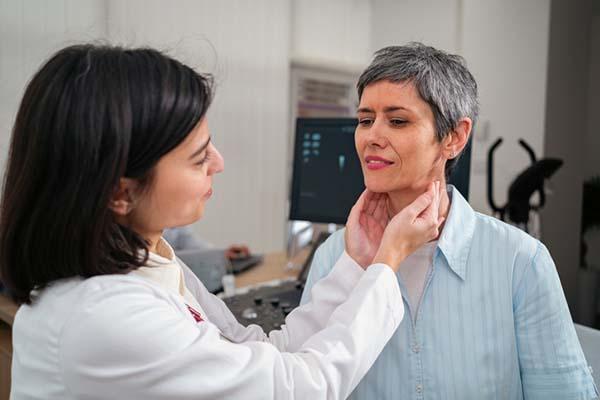 A female doctor using both hands to feel the thyroid gland of a mature female patient in an examination room.