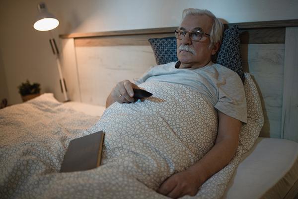 A senior man in bed, propped up by pillows and watching TV at night.