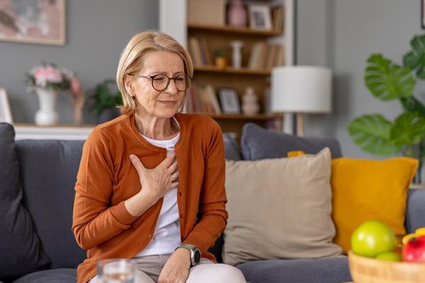 A mature woman holds a hand to her chest as she experiences chest pain while sitting on a couch in her home