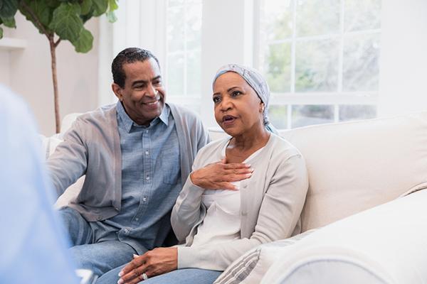 A mature woman holds a hand to her chest while describing symptoms; her husband sits next to her, listening and looking at her.