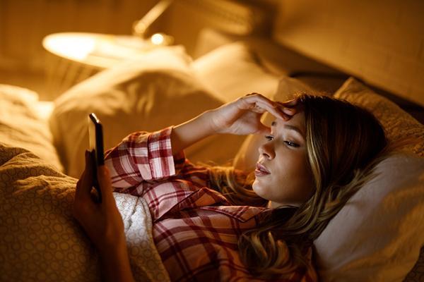 A young woman uses her smartphone while lying in bed in a dimly lit room.