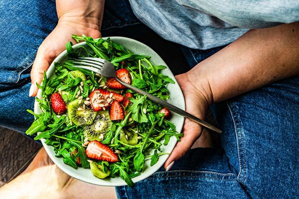 View from overhead of a man holding a bowl with salad containing greens, strawberries, kiwi,, sunflower seeds, and chia seeds.