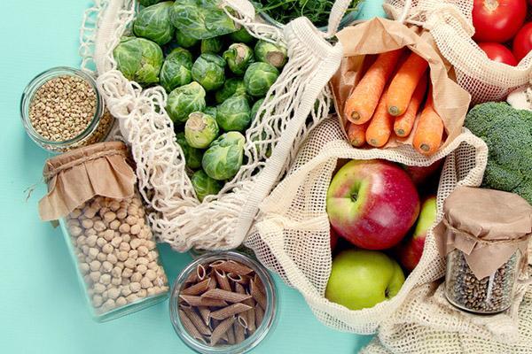 Mesh shopping bags and glass jars holding a variety of healthy foods: whole carrots, brussels sprouts, apples, tomatoes, whole-grain pasta, nuts, and seeds.