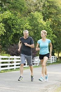 A man and a woman are seen running together on a rural road, with trees and a fence in the background.