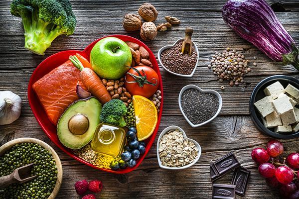 An assortment of heart-healthy foods displayed on a wood table: on a red heart-shaped plate are a granny smith apple, a salmon filet, carrot, broccoli, blueberries, tomato, olive oil, half an avocado, and an orange slice; next to the plate are small dishes with assorted nuts and seeds, and other foods scattered around them.