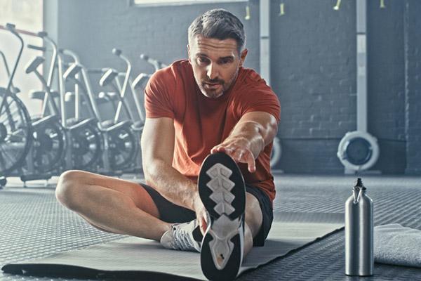 A mature man sits on the floor stretching one leg out in front of him after a workout in a gym.