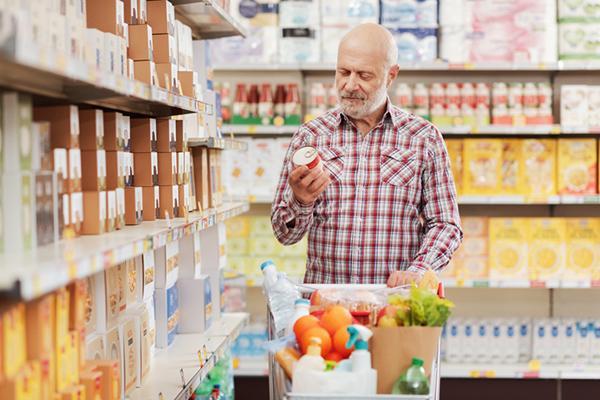A mature man holds a can and reads the product food label at the supermarket, standing behind a full cart in an aisle.