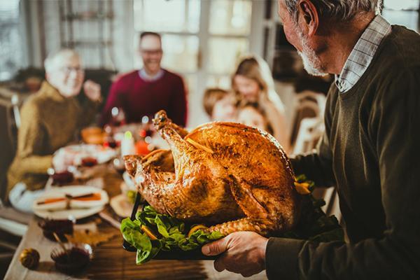 A senior man carries a platter with a large holiday turkey toward a table where other family members wait, out of focus.