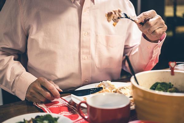 Cropped shot of the torso and arms of a man eating a meal; one hand holds a fork in front of his chest, the other holds a knife next to the plate.