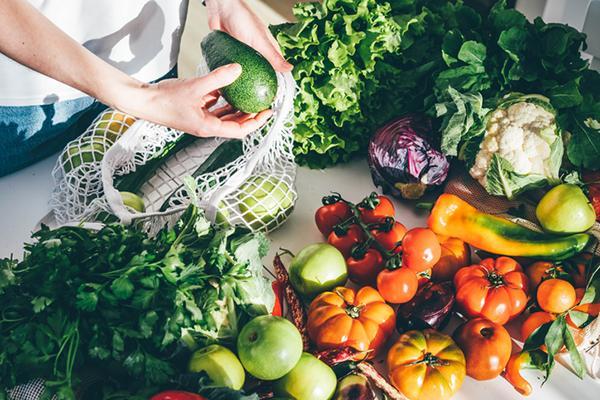 Cropped image of a woman unloading a large assortment of fresh produce from a net shopping bag onto a table.