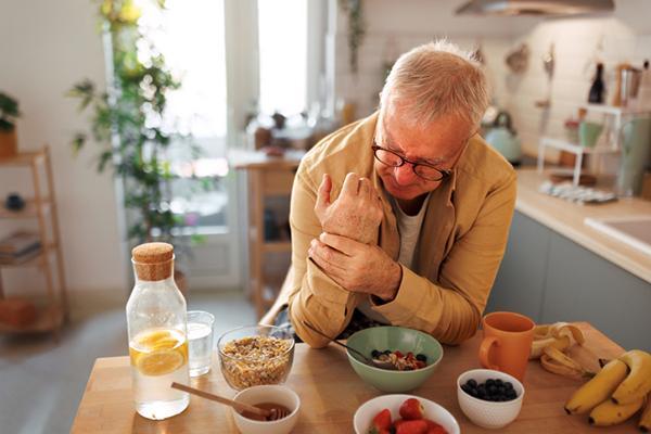 A senior man experiencing hand pain grasps his wrist with his other hand while having breakfast at home.