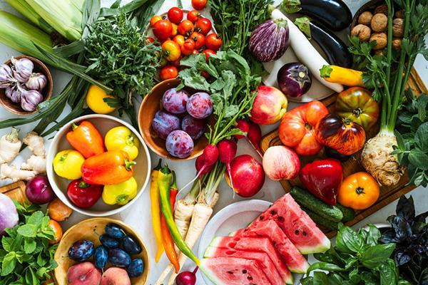 A view from above of an assortment of nutritious fruits and vegetables, including sliced watermelon, plums, grape tomatoes, peppers in assorted colors, apples, eggplants, turnips, and more.