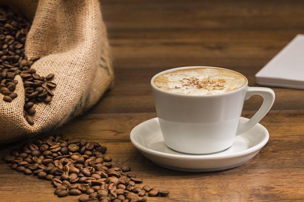 Close-up of a fresh cup of coffee next to a burlap sack of coffee beans with beans spilled out onto a wood table.