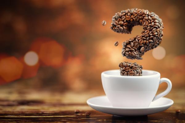 Coffee beans in the shape of a three-dmensional question mark hovering above a white coffee cup on a saucer.