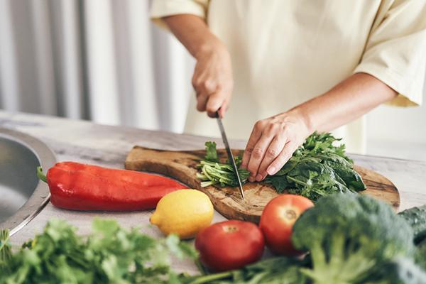 Close-up of hands chopping leafy greens on a cutting board, with a pepper, a lemon, and tomatoes on the counter in front of the board.