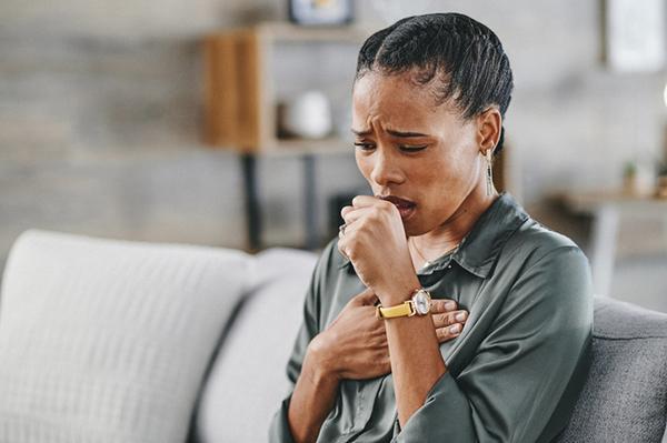 A coughing woman sitting on a sofa holds one hand to her chest and the other in front of her mouth.