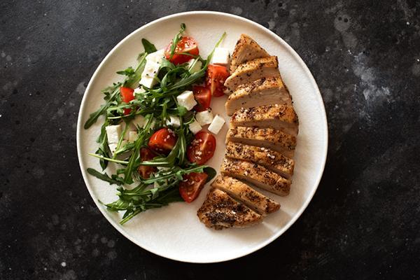 Overhead view of a plate with a serving of sliced grilled chicken breast and a salad with greens, tomatoes, and feta cheese.