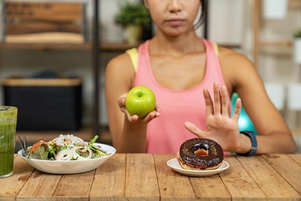 A woman holds a green apple and holds up her other hand, rejecting a tempting chocolate doughnut and opting for a nutritious salad and a green smoothie on a wooden table in front of her.