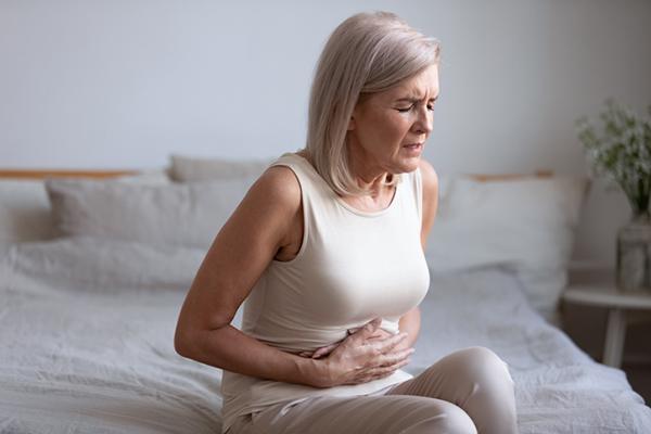 A woman sits on the edge of a bed, gripping her stomach with both hands and grimacing in pain.